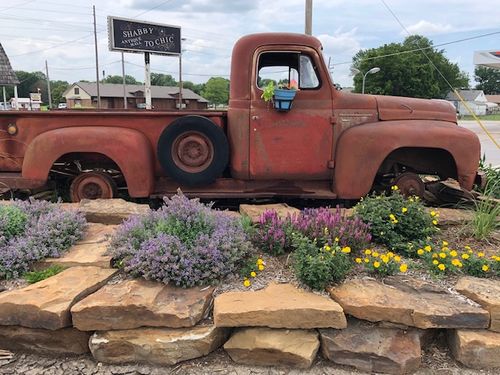 A rusty red vintage pickup truck sits on display behind a stone garden wall filled with purple and yellow flowers, possibly at a flea market near Coffeyville, KS.